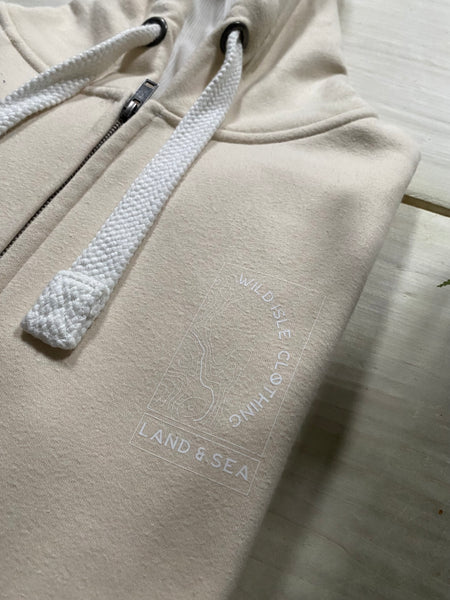 Beige Wild Isle Trail Zip Up hoodie with a logo and text on the chest, folded on a table