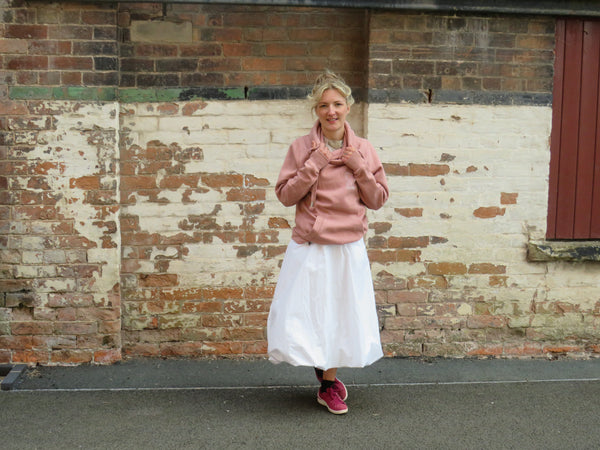 Woman wearing the wild isle clothing pink hoodie with white skirt and pink trainers against a brick wall.