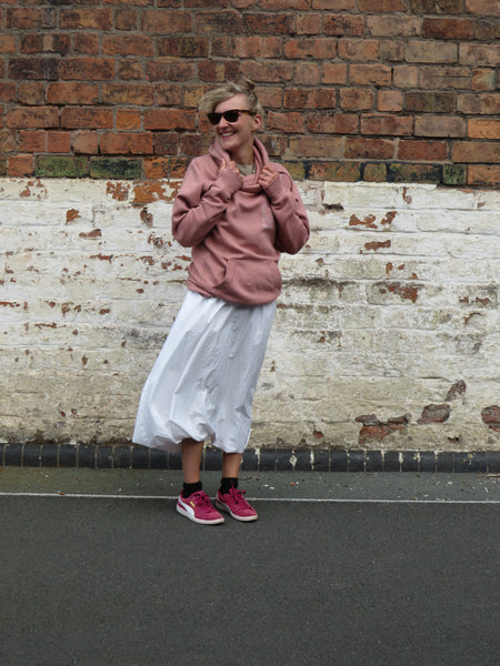 A woman wearing the wild isle hood in pink with a white skirt and pink pumps. against a brick wall.