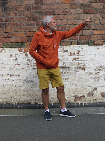 Man in the wild isle clothing copper beech hoodie and yellow shorts against a brick wall.