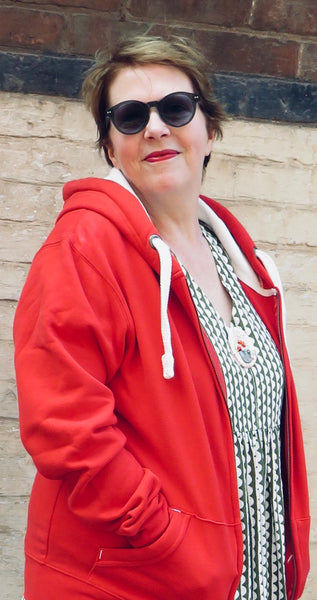 Person wearing a Wild Isle Trail Zip Up red hoodie and sunglasses against a brick wall.