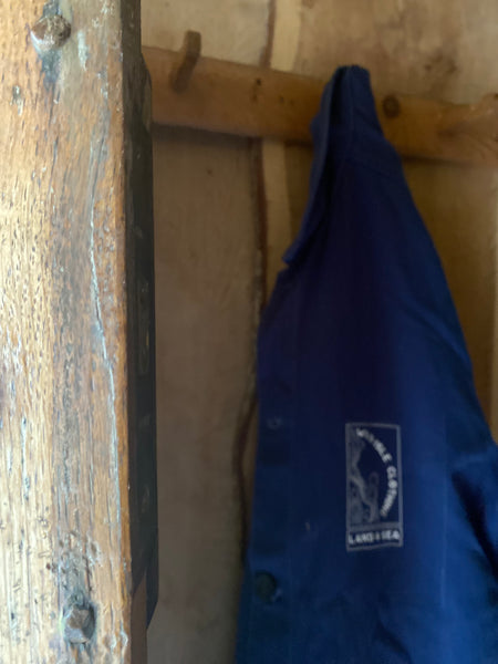 The wild isle crofters utility jacket in royal blue hanging on a wooden hook in the doorway of a cottage.