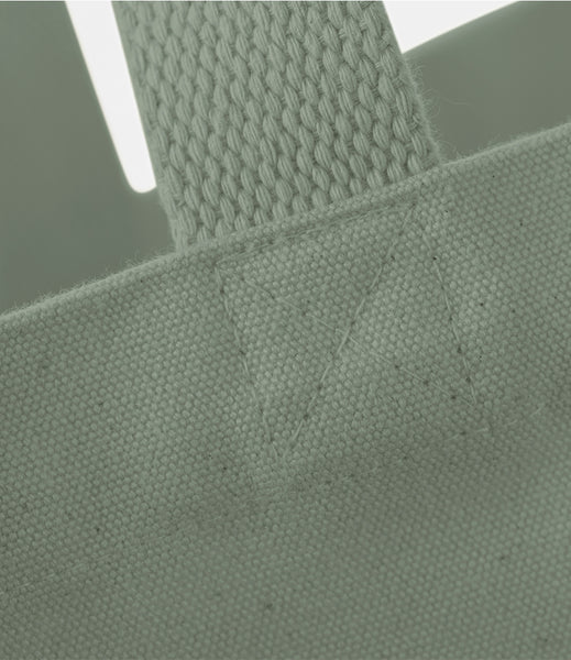 Close-up detail of a sage green canvas tote bag showing the woven strap attachment and fabric texture.