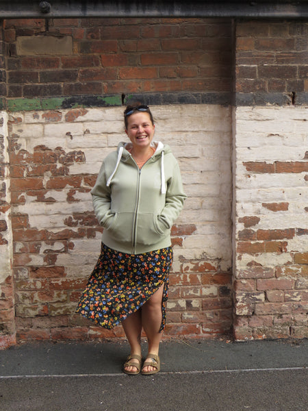 Woman standing against a textured brick wall wearing a wild isle clothing zip up hoodie in sea foam with logo
