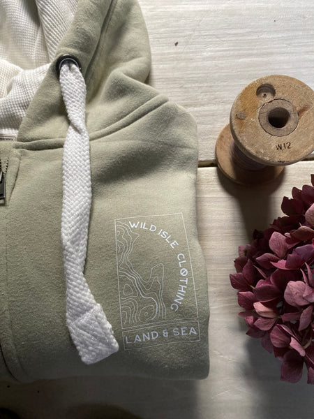 Green hoodie with 'Wild Isle Clothing' logo on a wooden surface with a spool and flowers.