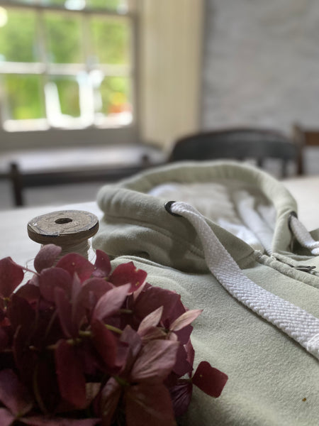 Spool of thread and bouquet of flowers and Sea Foam Wild Isle Clothing Hoodie on the table with a blurred background