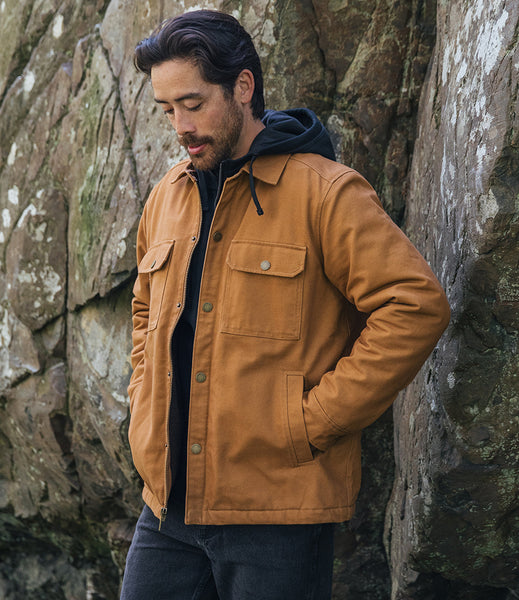 A man wearing a tan canvas utility jacket over a black hoodie and dark jeans, standing against a large rock wall.