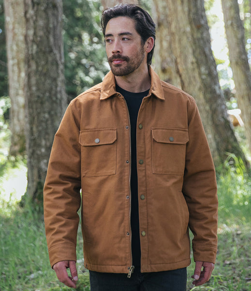 A man wearing a Tan canvas utility jacket over a black shirt, standing outdoors in a wooded area with trees in the background.