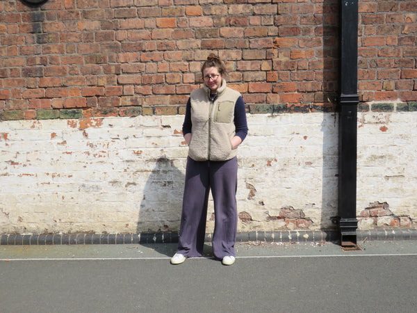 Person wearing a natural sand gilet and purple pants standing against a brick wall.