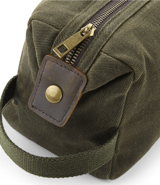 Wild Isle Waxed Canvas Wash bag with a dark brown leather strap tab and brass snap closure and carry strap next to a dark zipper with a brass pull.