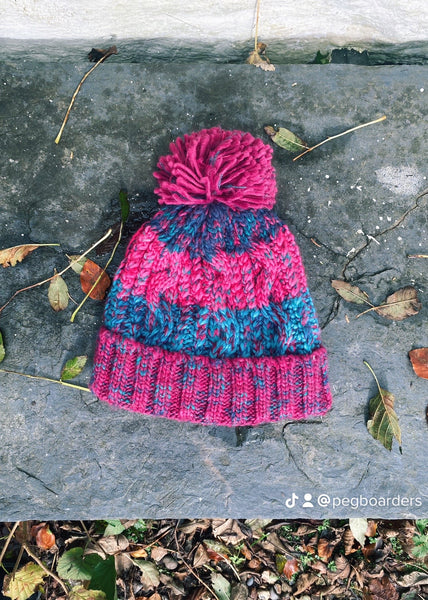 Pink and blue knitted beanie with a pom-pom on a stone bench with fallen leaves 